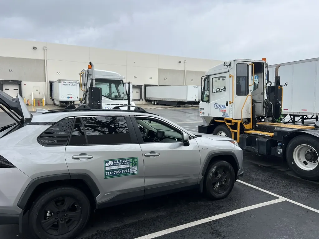 Clean Truck OC service vehicle side-on, with the CARB OBD/OVI credentialed-tester decal, beside a yard terminal tractor at a warehouse dock.